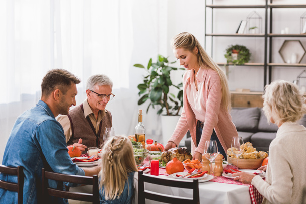 family sitting at table and mother holding plate with turkey in Thanksgiving day      - Photo, Image