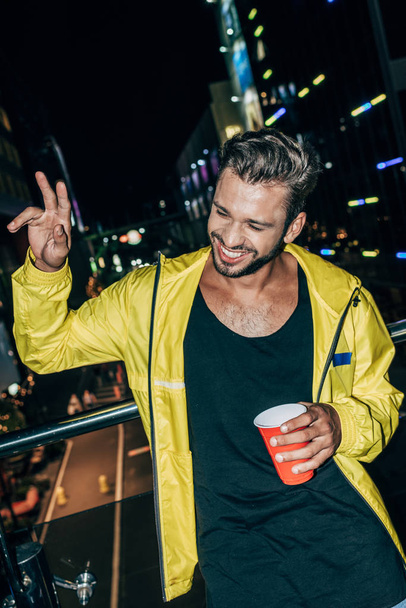 handsome man in yellow jacket showing peace sign and holding plastic cup in night city   - Fotografie, Obrázek