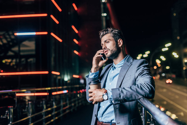 komea liikemies muodollinen kuluminen puhuu älypuhelimella ja pitämällä paperimuki yöllä kaupunki
   - Valokuva, kuva