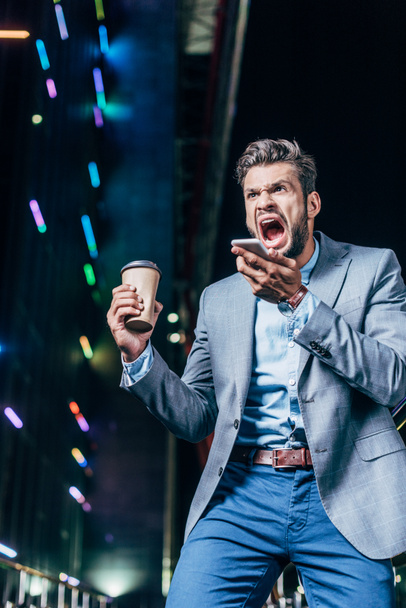 сердитий красивий бізнесмен в формальному одязі говорить на смартфоні і тримає паперовий стаканчик в нічному місті
   - Фото, зображення