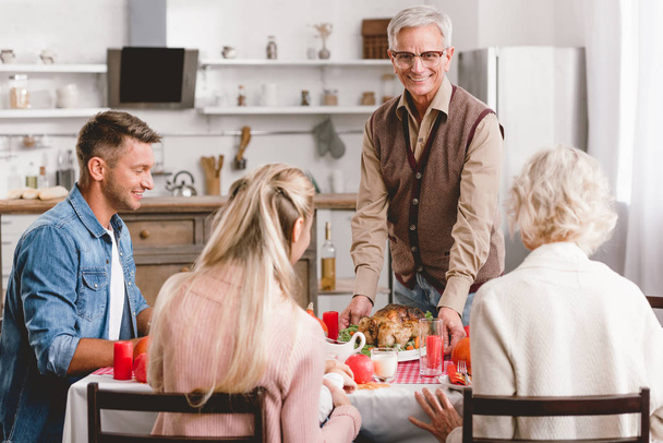 Familienmitglieder sitzen am Tisch und Großvater hält Teller mit Truthahn zum Erntedank       - Foto, Bild