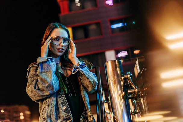 привлекательная женщина в джинсовой куртке и очках, смотрящая в камеру в ночном городе
  - Фото, изображение
