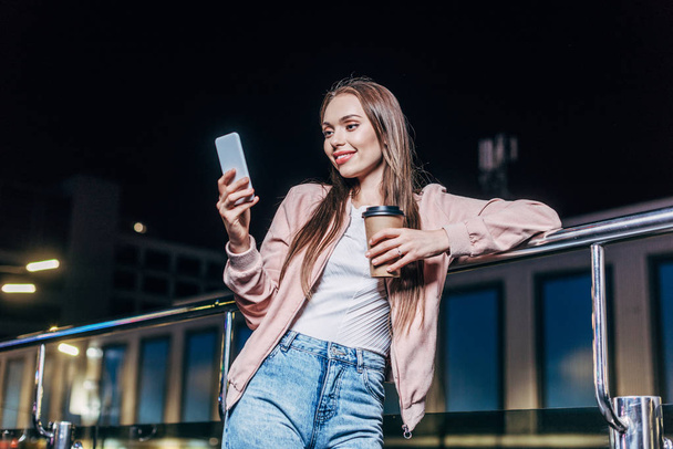 Lachende vrouw in roze jasje met behulp van smartphone en het houden van papier Cup in Night City  - Foto, afbeelding