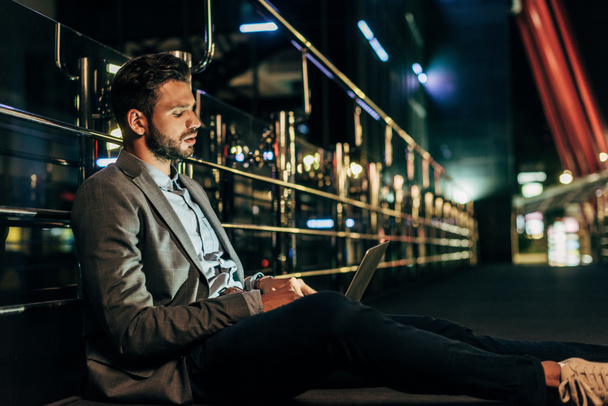 knappe zakenman in formele slijtage zittend en met behulp van laptop in Night City   - Foto, afbeelding