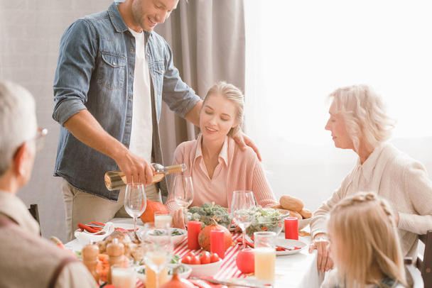 family members sitting at table and smiling father pouring wine in Thanksgiving day    - Photo, Image