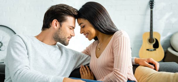 panoramic shot of african american woman and handsome man with closed eyes smiling and hugging in apartment  - Фото, изображение