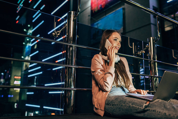 smiling woman in pink jacket with headphones talking on smartphone in night city  - Photo, Image