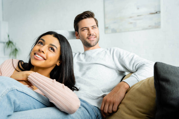 smiling african american woman lying on legs of handsome man in apartment  - 写真・画像
