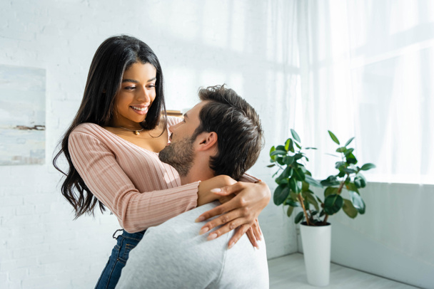 african american woman and handsome man smiling and hugging in apartment  - Foto, afbeelding