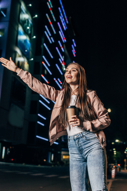 attractive woman in pink jacket hitching and holding paper cup in night city   - Fotografie, Obrázek