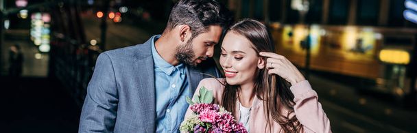 panoramic shot of handsome businessman giving bouquet and kissing attractive woman in night city  - Photo, Image
