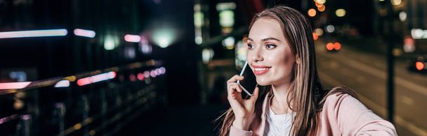 panoramic shot of attractive woman in pink jacket smiling and talking on smartphone in night city  - Photo, Image