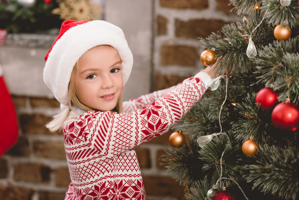 cute kid in santa hat and sweater decorating christmas tree - Photo, Image