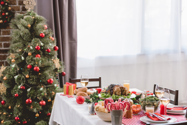 plate with tasty turkey, corn, candy canes, candles, gift and wine glasses on table and christmas tree - Foto, Imagen