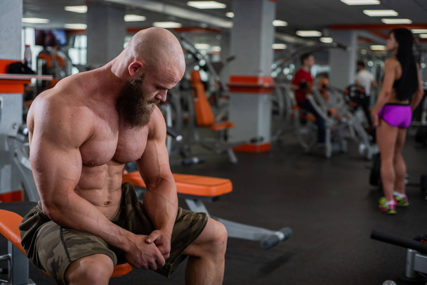 A young muscular guy is sitting on a bench in the gym and resting after a workout. Behind is a muscular dark-haired girl in shorts. Fitness center in the gym. - Foto, Imagem