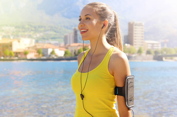 Happy fitness woman living a fit healthy lifestyle. Young girl wearing activewear and sports armband for phone and earphones, tech gear for running or cardio workout city lake on background. - Фото, зображення