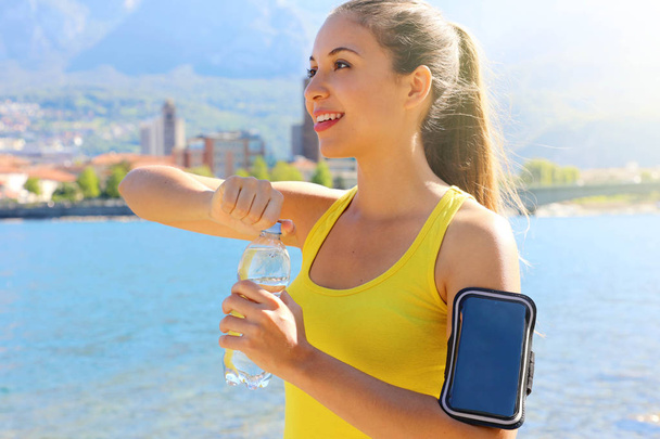 durstige Fitness-Frau öffnet Flasche Wasser nach dem Training im Freien. Fitte Frau nutzt Smartphone-Fitness-App auf Armband als Aktivitätstracker. - Foto, Bild