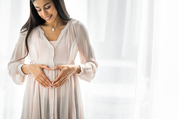 smiling pregnant african american woman showing heart sign in apartment  - Фото, изображение