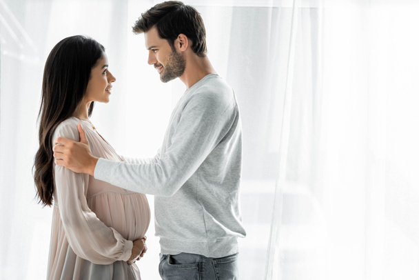 вид збоку красива і усміхнений чоловік обіймає вагітну афроамериканську жінку
  - Фото, зображення