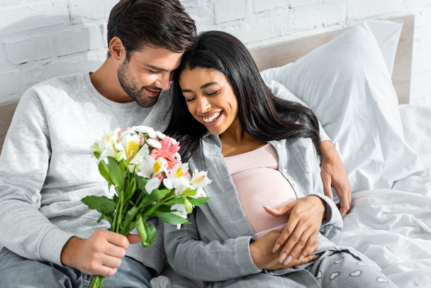 handsome man holding bouquet and hugging his smiling pregnant african american woman  - Foto, afbeelding