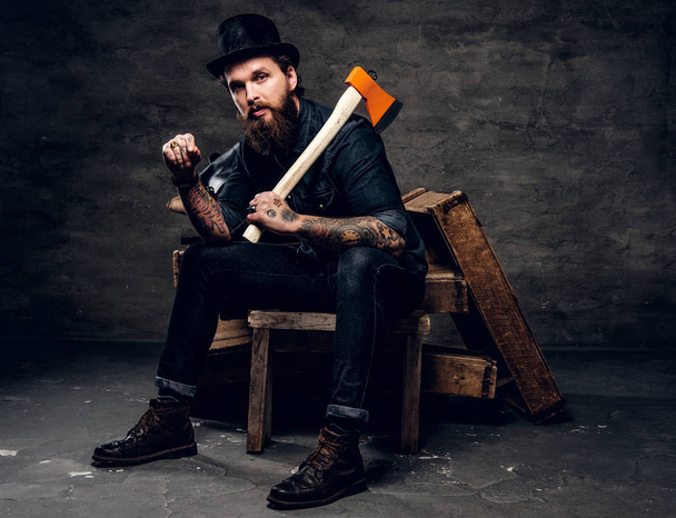 Serious bearded man in hat is sitting with axe in hands. - Photo, Image
