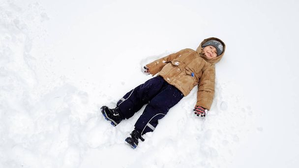 コートと灰色の帽子をかぶった笑顔の可愛い少年の肖像画をクローズアップし、降雪後の冬に雪の中で遊ぶのが楽しいです。公園の遊び場で雪のボールで遊ぶ - 写真・画像