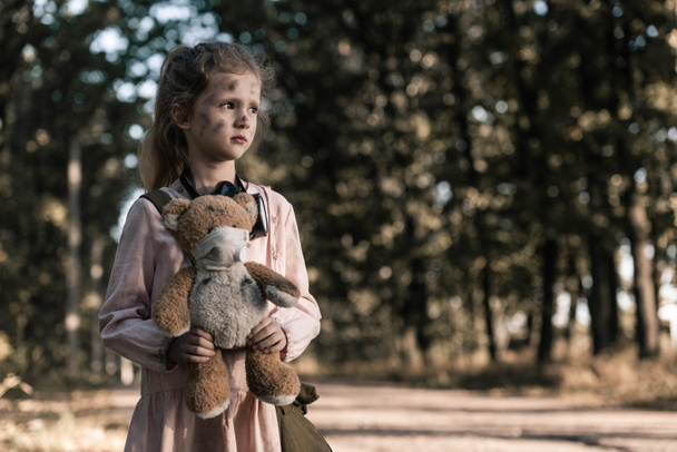 cute kid holding dirty teddy bear near trees in chernobyl, post apocalyptic concept - Photo, Image