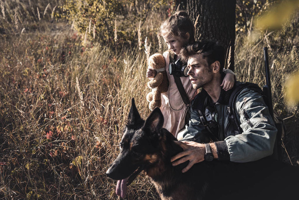 selective focus of man touching german shepherd dog near cute kid in field, post apocalyptic concept - Photo, Image