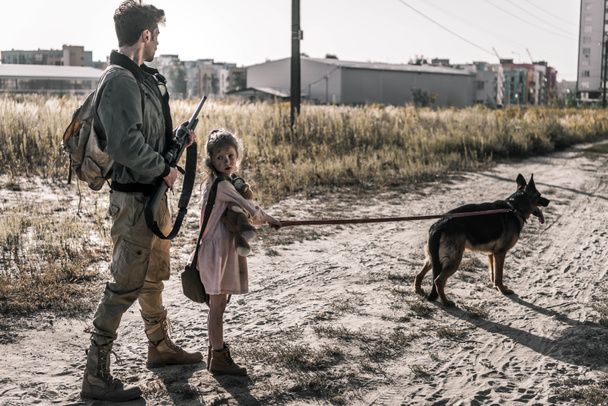 gewapende man met geweer in de buurt van kind met teddybeer en hond, post apocalyptisch concept - Foto, afbeelding
