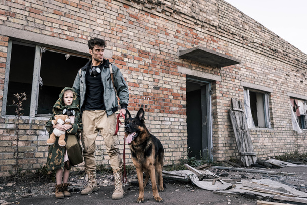 όμορφος άνδρας στέκεται με παιδί και γερμανικό τσοπανόσκυλο κοντά σε εγκαταλελειμμένο κτίριο, μετα-αποκαλυπτική έννοια - Φωτογραφία, εικόνα