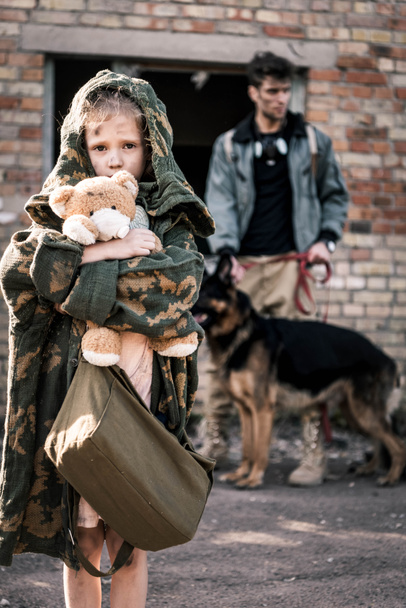 επιλεκτική εστίαση του παιδιού κρατώντας αρκουδάκι κοντά στον άνθρωπο με γερμανικό τσοπανόσκυλο κοντά σε εγκαταλελειμμένο κτίριο, μετα-αποκαλυπτική έννοια - Φωτογραφία, εικόνα