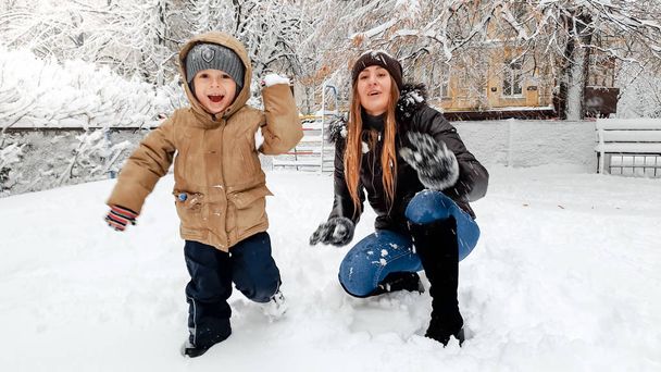 Primo piano colpo di allegra sorridente giovane madre in cappotto e cappello con il suo bel figlio in giacca beige divertirsi a giocare le palle di neve con la neve sul parco giochi nel parco
 - Foto, immagini