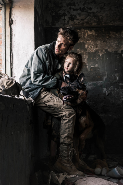 man knuffelen vuil kind in de buurt van duitse herder hond in verlaten gebouw, post apocalyptische concept - Foto, afbeelding