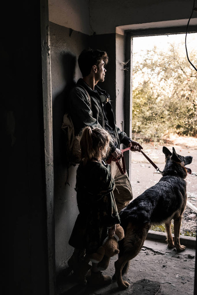man and kid with teddy bear standing near door with german shepherd dog, post apocalyptic concept - Foto, afbeelding