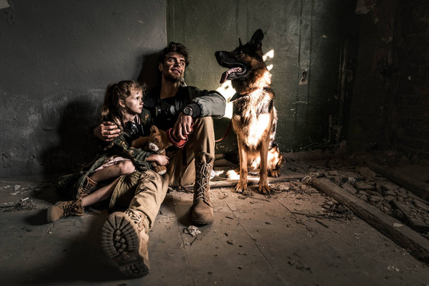 man and kid with teddy bear sitting on floor with german shepherd dog, post apocalyptic concept - Photo, Image