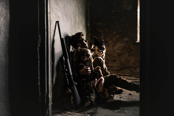 uomo e bambino in maschera antigas seduto sul pavimento vicino pistola e cane pastore tedesco, concetto post apocalittico
 - Foto, immagini