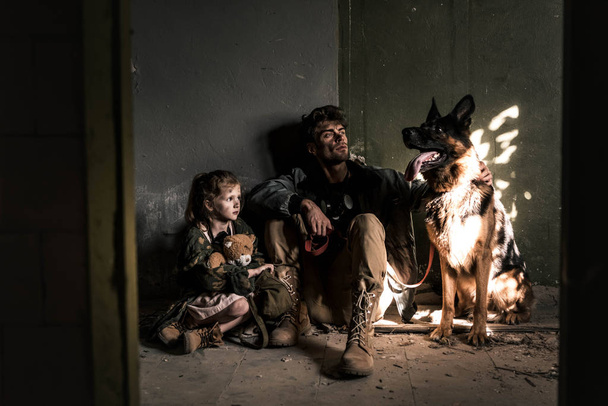 hombre guapo y niño con oso de peluche sentado en el suelo con perro pastor alemán, concepto post apocalíptico
 - Foto, Imagen