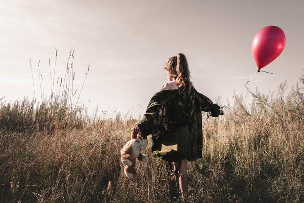 back view of kid with teddy bear holding balloon in field, post apocalyptic concept - Photo, Image