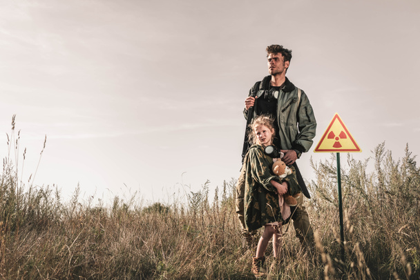 handsome man with gun near cute kid with teddy bear and toxic symbol, post apocalyptic concept - Photo, Image