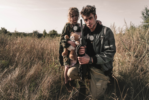 hombre guapo sosteniendo pistola cerca de niño lindo con juguete suave en el campo, concepto post apocalíptico
 - Foto, Imagen