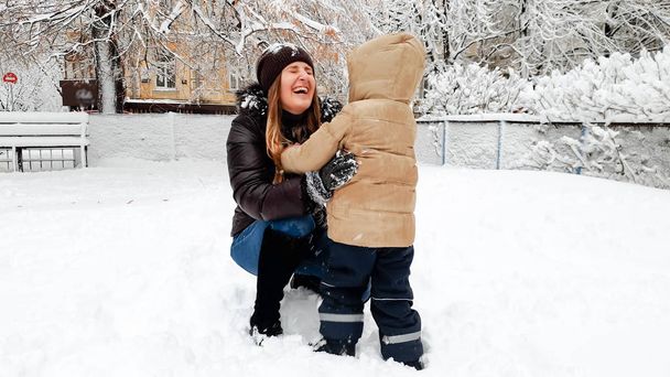ベージュのジャケットで彼女のかわいい息子とコートで笑顔の母の写真は、公園の遊び場で雪のボールを再生する楽しみを持っています - 写真・画像