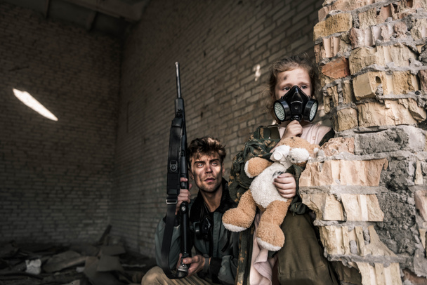 Kind in Gasmaske hält Teddybär neben gutaussehendem Mann mit Waffe, postapokalyptisches Konzept - Foto, Bild
