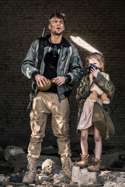 pohledný muž drží baňku poblíž dítěte v plynové masce, post apokalyptický koncept - Fotografie, Obrázek