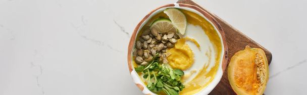 top view of autumnal mashed pumpkin soup in bowl on wooden cutting board with pumpkin on marble surface, panoramic shot - Fotó, kép