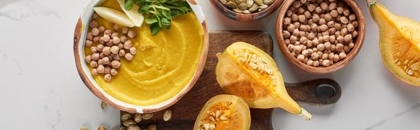 top view of autumnal mashed pumpkin soup in bowl on wooden cutting board near pumpkin, chickpea on marble surface, panoramic shot - Photo, Image