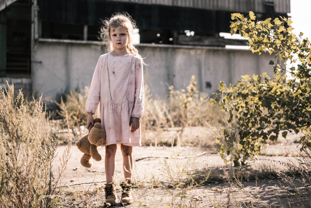 ребенок с грязным лицом, смотрящий в камеру, держа плюшевого мишку, пост апокалиптической концепции
 - Фото, изображение
