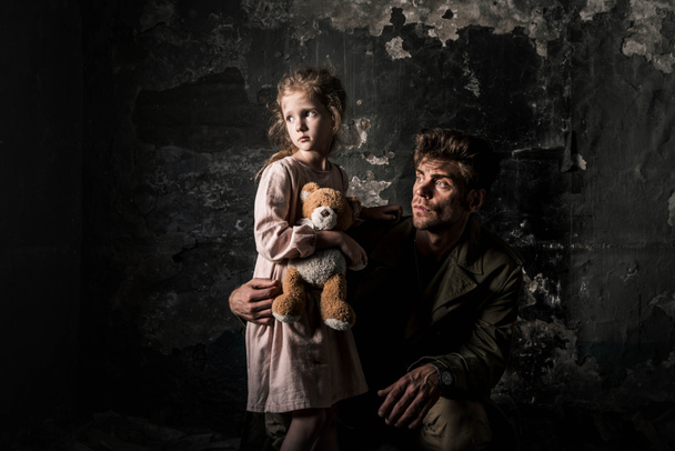 boos man zitten in de buurt van kind met teddybeer in vuile kamer, post apocalyptische concept - Foto, afbeelding