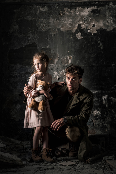 schöner Mann sitzt neben aufgebrachtem Kind mit Teddybär im schmutzigen Raum, postapokalyptisches Konzept - Foto, Bild