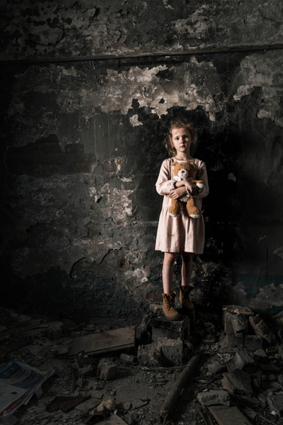 Üzgün çocuk ayakta duruyor ve oyuncak ayıyı kirli odada tutuyor, kıyamet sonrası konsepti - Fotoğraf, Görsel