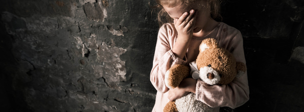 Panoramaaufnahme eines aufgebrachten Kindes, das das Gesicht berührt, während es einen Teddybär in einem schmutzigen Raum hält, postapokalyptisches Konzept - Foto, Bild